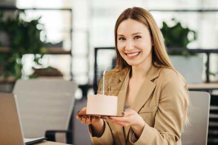 Photo for Happy businesswoman holding cake with lightened candle in the office and looking at camera. - Royalty Free Image