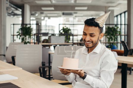 Foto de Feliz empresario hindú sosteniendo pastel con vela aligerada en la oficina y mirando a la cámara. - Imagen libre de derechos