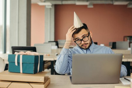 Photo for After birthday party. Bearded office worker in cone hat sitting at workplace with laptop and gift with festive ribbon. - Royalty Free Image