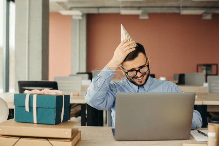 Photo for Happy positive bearded office worker in cone hat sitting at workplace with laptop and gift with festive ribbon. - Royalty Free Image