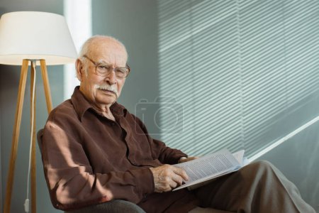 Téléchargez les photos : Homme âgé assis seul sur un fauteuil dans le salon portant des lunettes et tenant un livre, regardant attentivement la caméra - en image libre de droit
