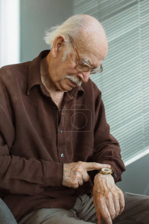 Téléchargez les photos : Un vieil homme solitaire dans le salon regarde sa montre au poignet. En attendant la visite d'un ami ou de parents. - en image libre de droit