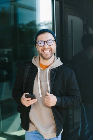 Photo for Positive young guy holding smartphone in his hands, looking at camera standing on street. Bearded man in eyeglass wears casual clothes. Cell phone usage concept - Royalty Free Image