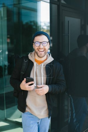Foto de Positive young guy holding smartphone in his hands, looking at camera standing on street. Bearded man in eyeglass wears casual clothes. Cell phone usage concept - Imagen libre de derechos