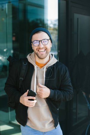 Photo for Positive young guy holding smartphone in his hands, looking at camera standing on street. Bearded man in eyeglass wears casual clothes. Cell phone usage concept - Royalty Free Image