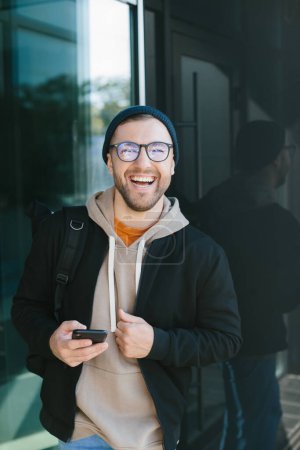 Foto de Positive young guy holding smartphone in his hands, looking at camera standing on street. Bearded man in eyeglass wears casual clothes. Cell phone usage concept - Imagen libre de derechos
