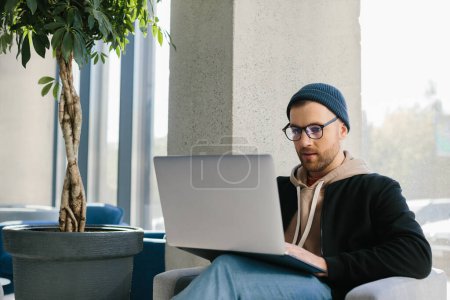 Photo for Young handsome freelancer guy working with laptop in office space. A man in glasses and a hat is holding a laptop while sitting in a chair in the hall. - Royalty Free Image