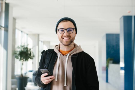 Foto de A positive young man with a smartphone in his hands is looking at the camera. A young man in glasses and a cap is standing in an office space. - Imagen libre de derechos