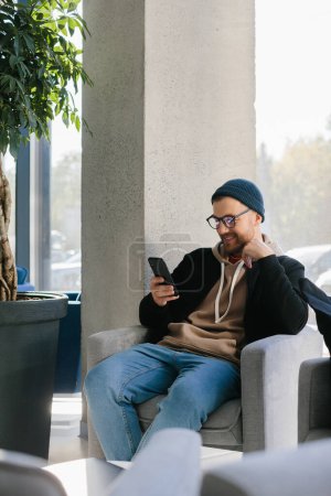 Téléchargez les photos : A young handsome guy with a phone in his hands is waiting for a job interview in an office space. A man in glasses and a cap is holding a phone while sitting in a chair in the hall. - en image libre de droit