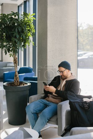 Photo for A young handsome guy with a phone in his hands is waiting for a job interview in an office space. A man in glasses and a cap is holding a phone while sitting in a chair in the hall. - Royalty Free Image