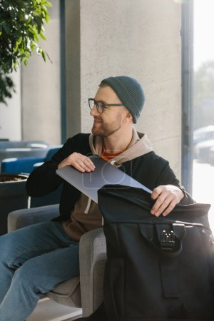 Foto de Young handsome freelancer guy working with laptop in office space. A man in glasses and a hat is holding a laptop while sitting in a chair in the hall. - Imagen libre de derechos