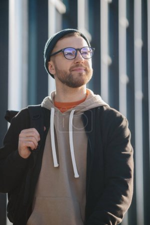 Photo for A young man with a backpack and casual clothes on the background of an office building. - Royalty Free Image