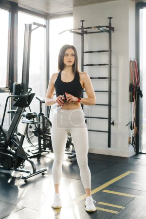 Photo for Athlete girl in leggings and a top with a bottle of water in the gym. The girl is engaged in fitness in the gym. Leggings mockup. - Royalty Free Image