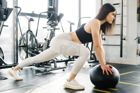 Photo for Young caucasian woman in black topic and white leggins do exercises with black fitness ball in gy - Royalty Free Image