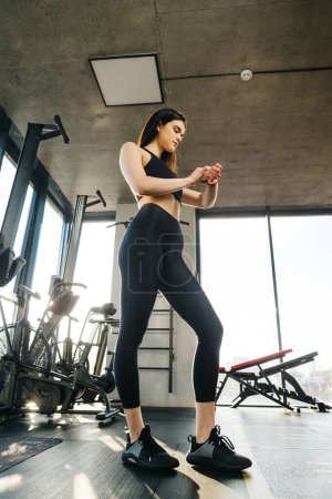 Téléchargez les photos : An athletic girl wearing black leggings and a top adjusts her smart watch in the gym. - en image libre de droit