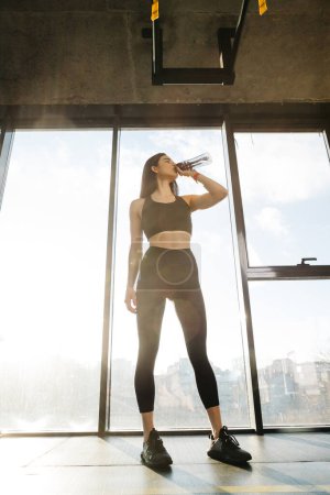 Foto de Attractive sporty girl drinks water while standing by the window in a fitness class. - Imagen libre de derechos