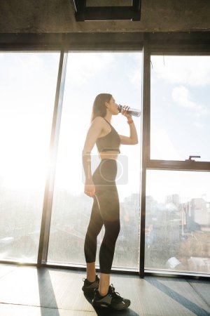 Photo for Attractive sporty girl drinks water while standing by the window in a fitness class. - Royalty Free Image
