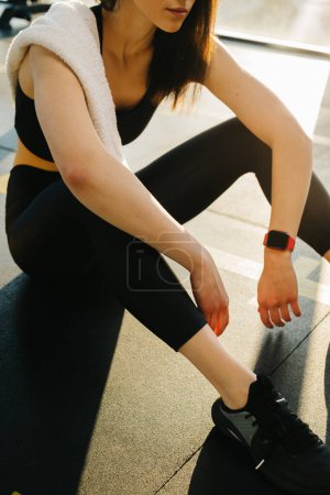 Photo for A young woman in black leggings and a top with a towel on her shoulder rests between exercises while sitting on the floor in the gym. - Royalty Free Image