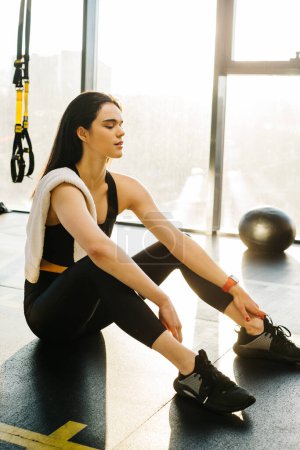 Téléchargez les photos : A young woman in black leggings and a top with a towel on her shoulder rests between exercises while sitting on the floor in the gym. - en image libre de droit