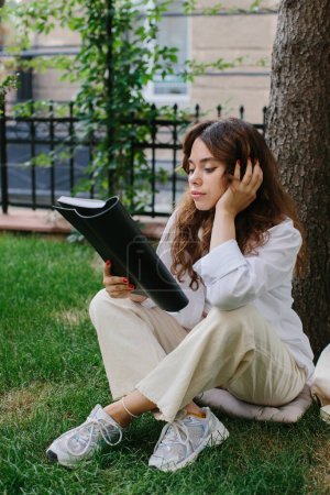 Téléchargez les photos : Modèle d'image de magazine ou livre. La fille se détend sur la pelouse dans la cour du café, lit un livre. - en image libre de droit