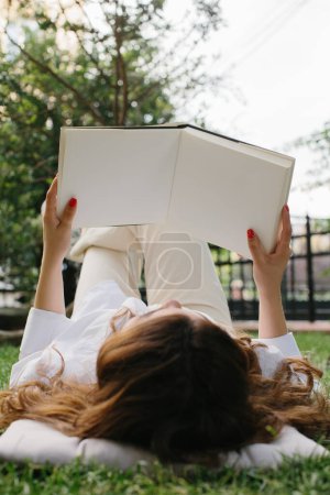 Téléchargez les photos : Magazine or book image mockup. A young brunette girl reads a book lying on the lawn in the summer garden - en image libre de droit