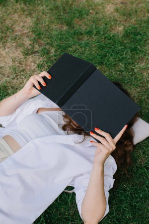 Téléchargez les photos : Modèle d'image de magazine ou livre. La fille se détend sur la pelouse dans la cour du café, lit un livre. - en image libre de droit