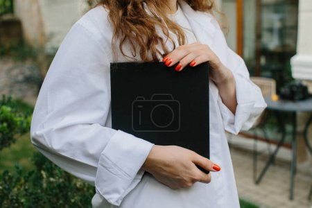 Photo for Magazine or book image mockup. Young attractive girl holding a closed book in her hands while standing in the yard. - Royalty Free Image