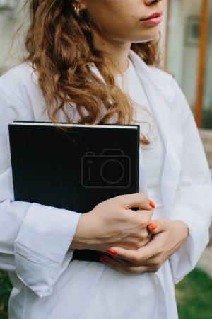 Foto de Magazine or book image mockup. Young attractive girl holding a closed book in her hands while standing in the yard. - Imagen libre de derechos
