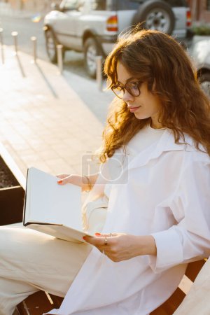 Foto de Magazine or book image mockup. A girl reads a book sitting on a bench on a city street at sunset. - Imagen libre de derechos
