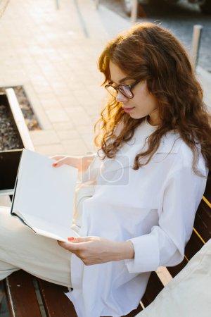 Téléchargez les photos : Magazine or book image mockup. A girl reads a book sitting on a bench on a city street at sunset. - en image libre de droit