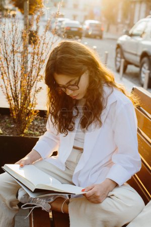 Téléchargez les photos : Magazine or book image mockup. A girl reads a book sitting on a bench on a city street at sunset. - en image libre de droit