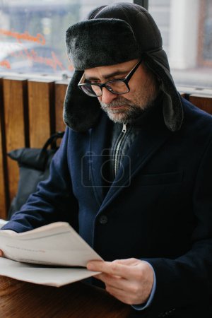 Photo for A gray-haired man in warm clothes reads a fresh newspaper and drinks hot tea or coffee on the street. - Royalty Free Image