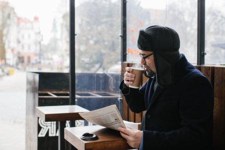 Téléchargez les photos : Un homme aux cheveux gris en vêtements chauds lit un journal frais et boit du thé ou du café chaud dans la rue. - en image libre de droit