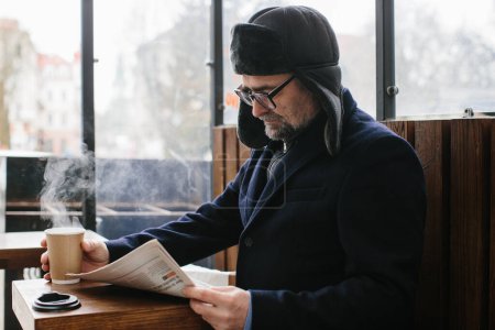 Téléchargez les photos : Un homme aux cheveux gris en vêtements chauds lit un journal frais et boit du thé ou du café chaud dans la rue. - en image libre de droit