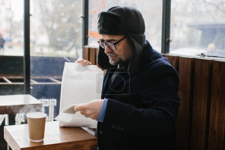 Téléchargez les photos : Un homme aux cheveux gris en vêtements chauds va déjeuner dans une fast-food de rue. - en image libre de droit