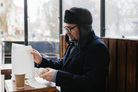 Téléchargez les photos : Un homme aux cheveux gris en vêtements chauds va déjeuner dans une fast-food de rue. - en image libre de droit
