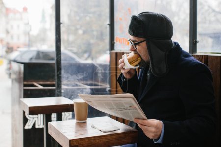 Téléchargez les photos : Un barbu d'âge moyen mange un délicieux hamburger et lit le journal d'aujourd'hui dans une rue de la ville en hiver. Concept de nourriture de rue. - en image libre de droit