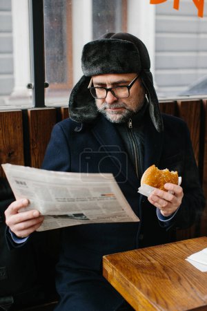 Téléchargez les photos : Un barbu d'âge moyen mange un délicieux hamburger et lit le journal d'aujourd'hui dans une rue de la ville en hiver. Concept de nourriture de rue. - en image libre de droit
