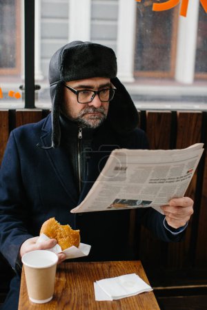 Photo for A middle-aged bearded man eats a tasty burger and reads today's newspaper on a city street in winter. Street food concept. - Royalty Free Image