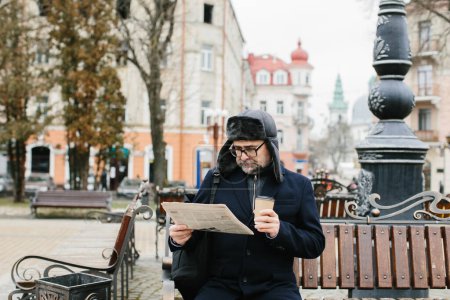 Téléchargez les photos : Un homme âgé profite d'une froide matinée d'hiver dans la ville. Un homme s'assoit sur un banc sur la place, lit un journal et boit du café. - en image libre de droit