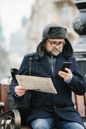 Téléchargez les photos : Un homme âgé profite d'une froide matinée d'hiver dans la ville. Un homme s'assoit sur un banc sur la place, lit un journal et boit du café. - en image libre de droit
