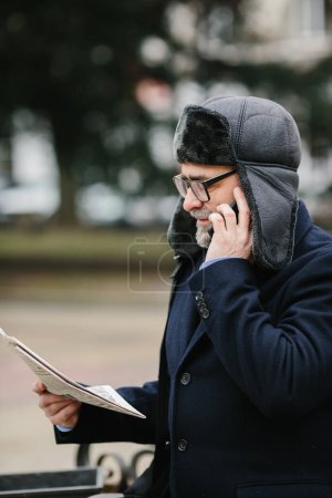 Téléchargez les photos : An elderly man enjoys a cold winter morning in the city. A man sits on a bench in the square, reads a newspaper and cheerfully chats with friends on a mobile phone. - en image libre de droit