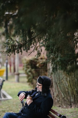 Téléchargez les photos : Un homme âgé profite d'une froide matinée d'hiver dans la ville. Un homme assis sur un banc sur la place, le concept de paix et de vitalité. - en image libre de droit