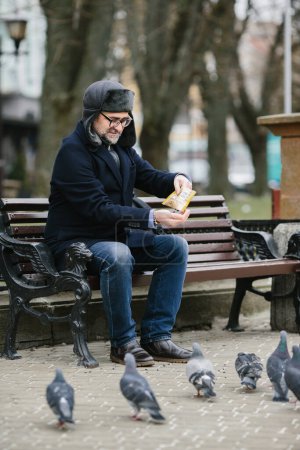 Photo for An elderly man enjoys a cold winter morning in the city. A gray-haired man feeds pigeons while sitting on a bench in the square. - Royalty Free Image