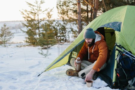 Téléchargez les photos : Un jeune homme barbu se repose dans les montagnes d'hiver près d'une tente. Un homme-voyageur avec une barbe dans une casquette et une veste chaude se réchauffe en buvant du thé ou du café chaud après une randonnée. Voyages, mode de vie - en image libre de droit