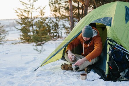 Téléchargez les photos : Un jeune homme barbu se repose dans les montagnes d'hiver près d'une tente. Un homme-voyageur avec une barbe dans une casquette et une veste chaude se réchauffe en buvant du thé ou du café chaud après une randonnée. Voyages, mode de vie - en image libre de droit