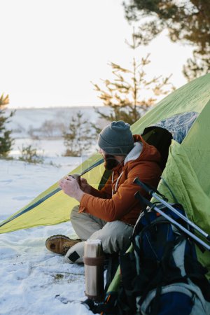 Photo for A young bearded man rests in the winter mountains near a tent. A man-traveler with a beard in a cap and a warm jacket warms up by drinking hot tea or coffee after a hike. Travel, lifestyle - Royalty Free Image