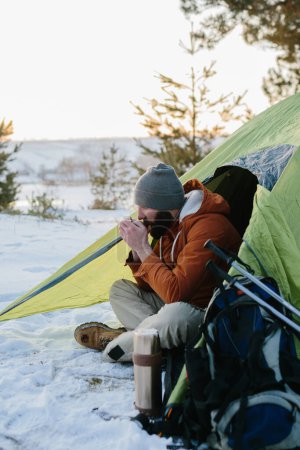 Photo for A young bearded man rests in the winter mountains near a tent. A man-traveler with a beard in a cap and a warm jacket warms up by drinking hot tea or coffee after a hike. Travel, lifestyle - Royalty Free Image