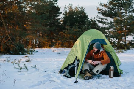 Téléchargez les photos : Un touriste se repose après une randonnée, assis près d'une tente avec une tasse de café, dans les rayons chauds du soleil couchant dans une forêt d'hiver. - en image libre de droit
