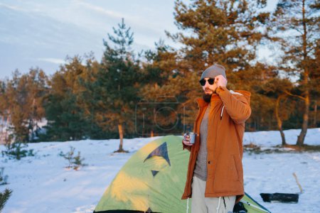 Téléchargez les photos : Un touriste, debout près de la tente avec une tasse de thé ou de café, profite du coucher de soleil dans la forêt d'hiver. - en image libre de droit